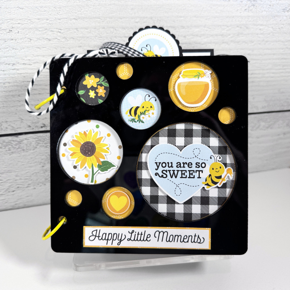 Happy Little Moments Scrapbook Album with bees, flowers, honey, and circle cutouts displayed on a clear acrylic stand.